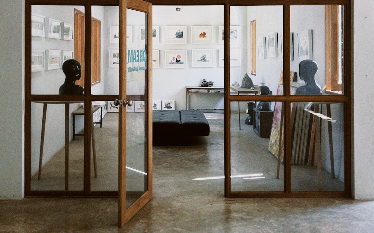 A bright gallery space hung with framed prints, seen through glass doors.